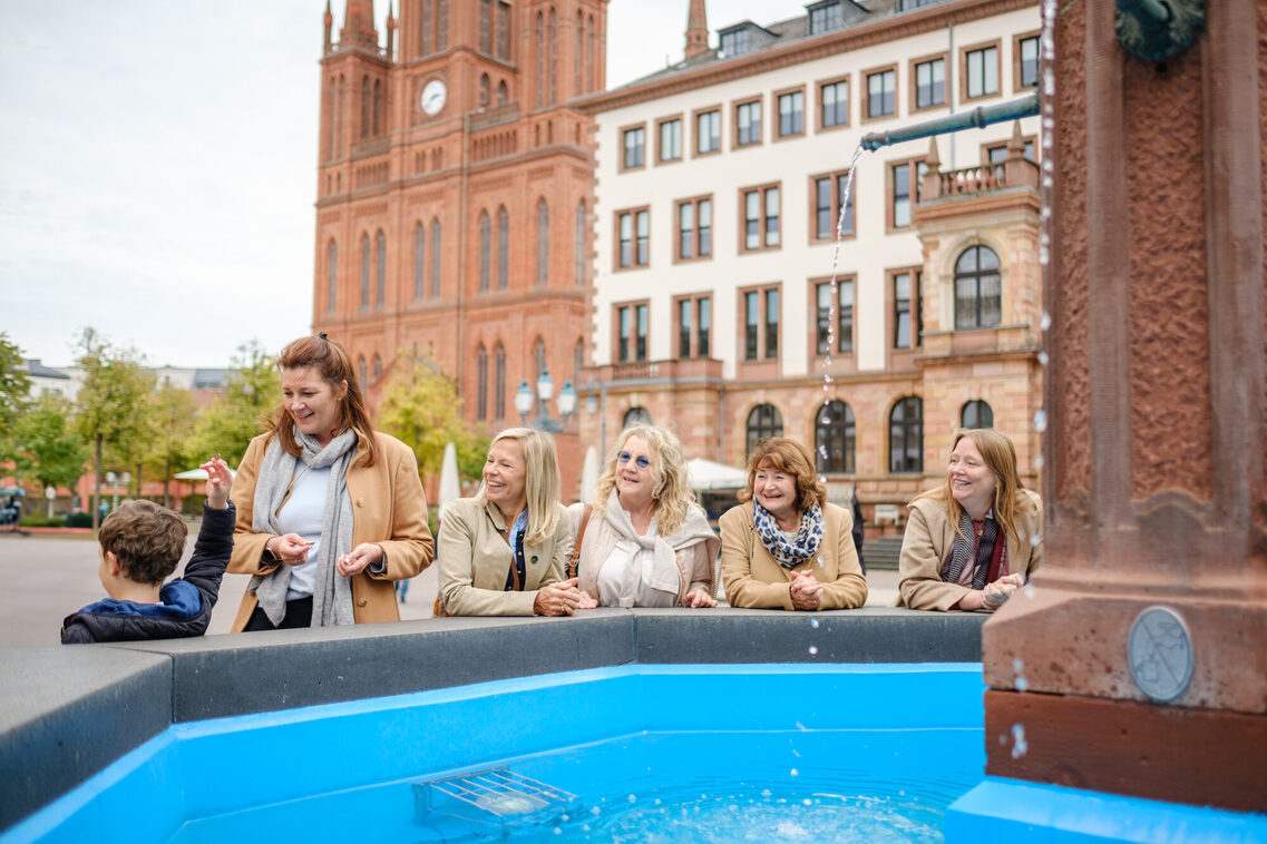 Fünf Frauen und ein kleiner Junge stehen vor einem Brunnen mit Wasser