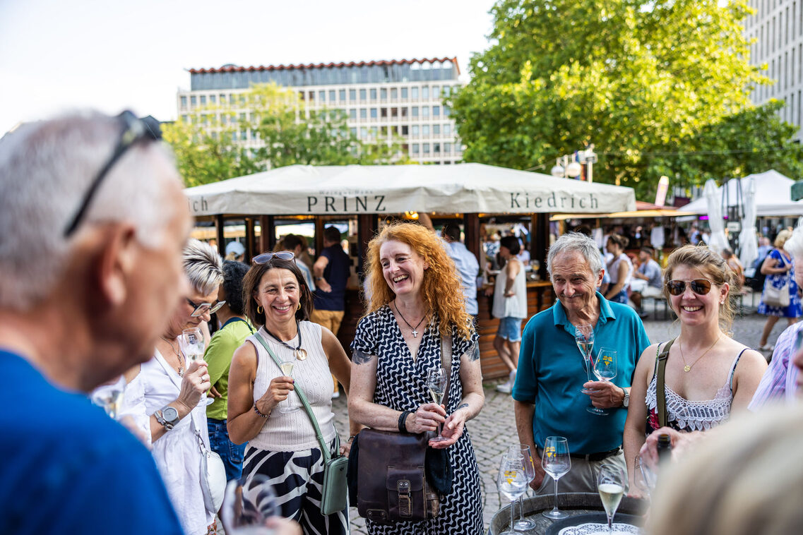 Frauen und Männer stehen vor Weinstand und lachen