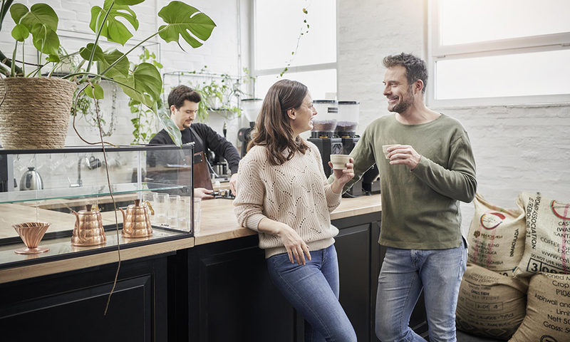 Zwei Menschen unterhalten sich mit Kaffe in der Hand an eine Theke gelehnt, Kaffeesäcke im Hintergrund.