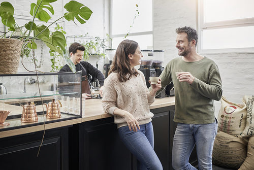 Zwei Menschen unterhalten sich mit Kaffe in der Hand an eine Theke gelehnt, Kaffeesäcke im Hintergrund.