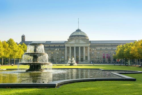 Kurhaus Wiesbaden mit von Bäumen gesäumtem Bowling Green mit zwei Brunnen.